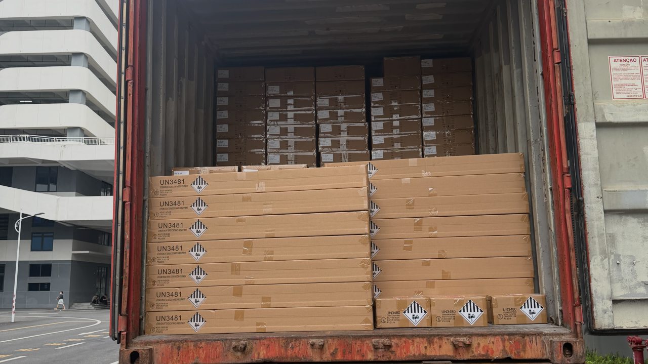 Stacked cardboard boxes with UN3481 lithium ion battery warning labels loaded inside a shipping container for transport.